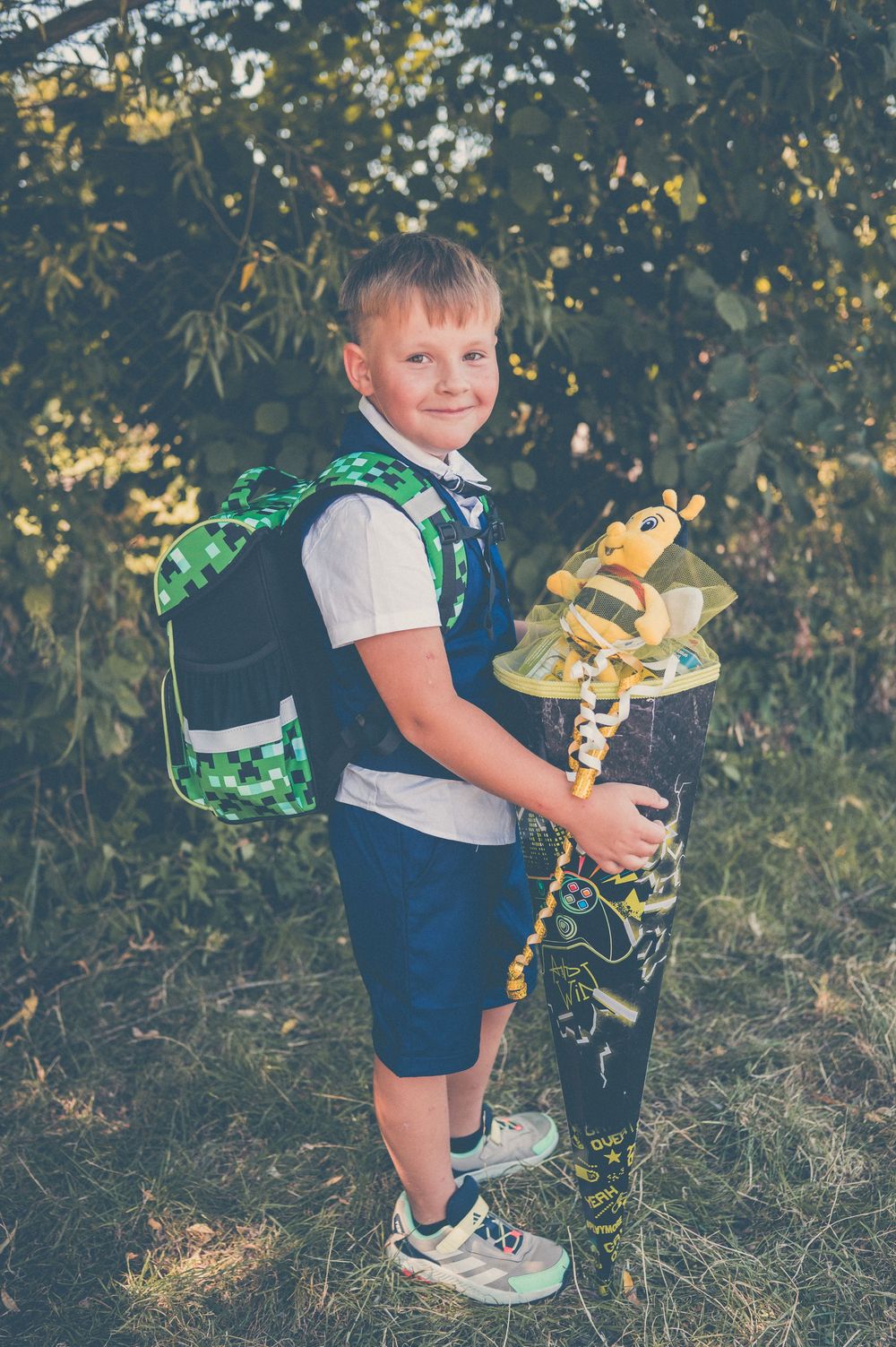 A smiling boy with a Minecraft backpack holds a Schultüte with a bee toy, standing outdoors on his first day of school.