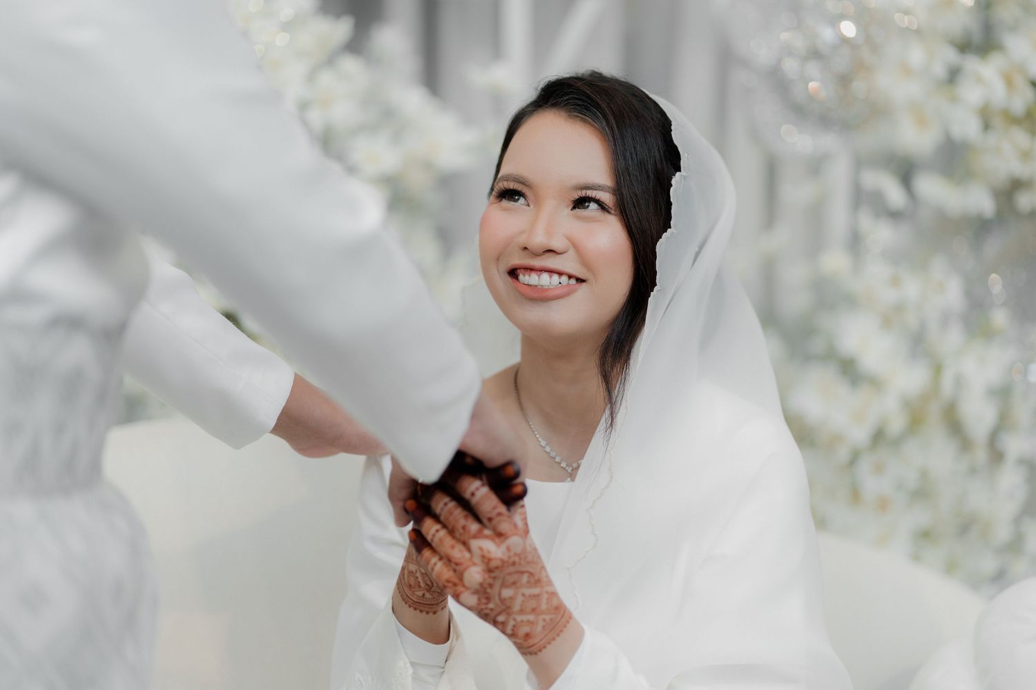 Bride in white gown and groom in suit share a joyful hug with a guest at a waterfront wedding ceremony at dusk.