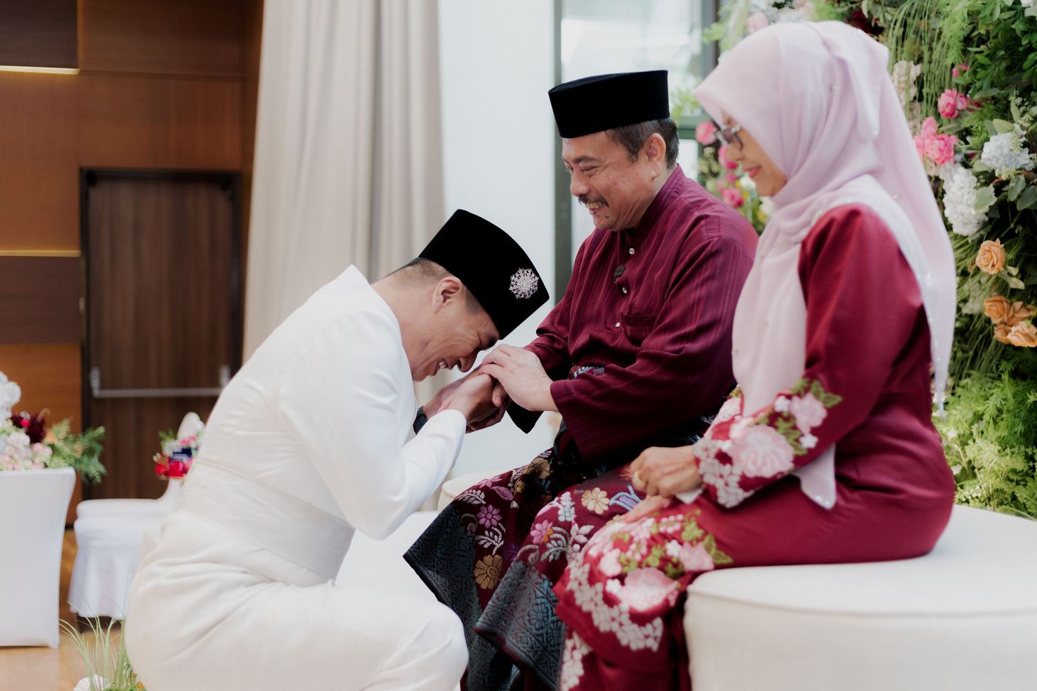 Traditional Malay wedding ceremony with couple seated on stage, performers in green, floral decor, and waterfront backdrop.