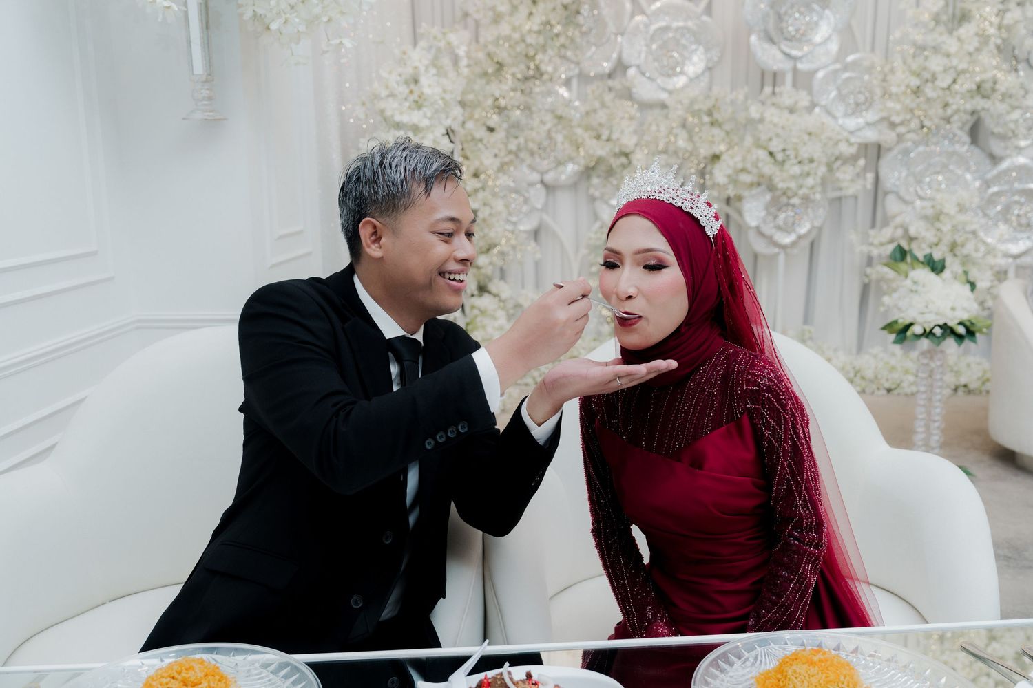Traditional Malay wedding ceremony with couple seated on stage, performers in green, floral decor, and waterfront backdrop.