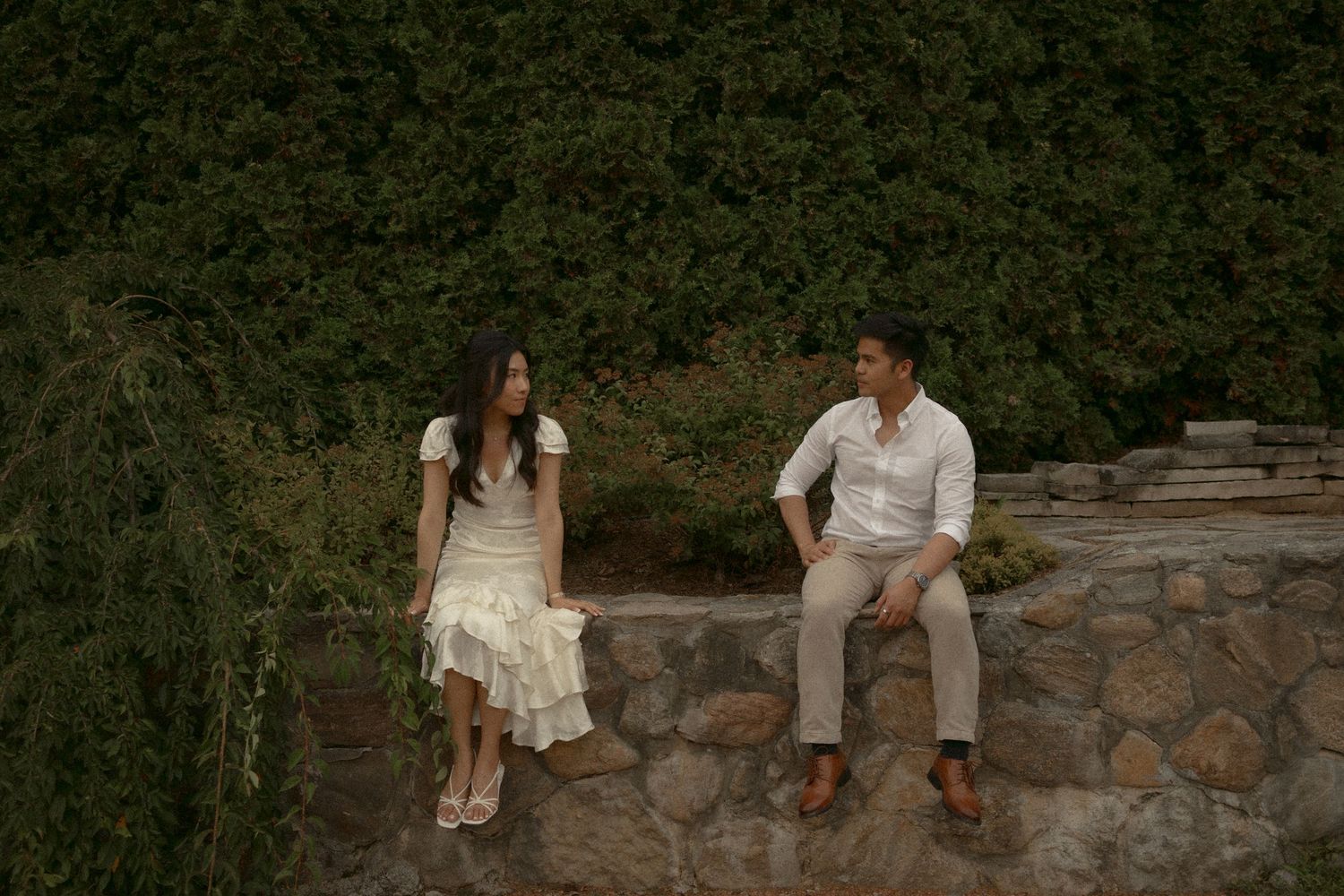 A couple sits apart on a stone wall surrounded by lush green hedges in a romantic outdoor garden setting. Whistling Gardens Ontario