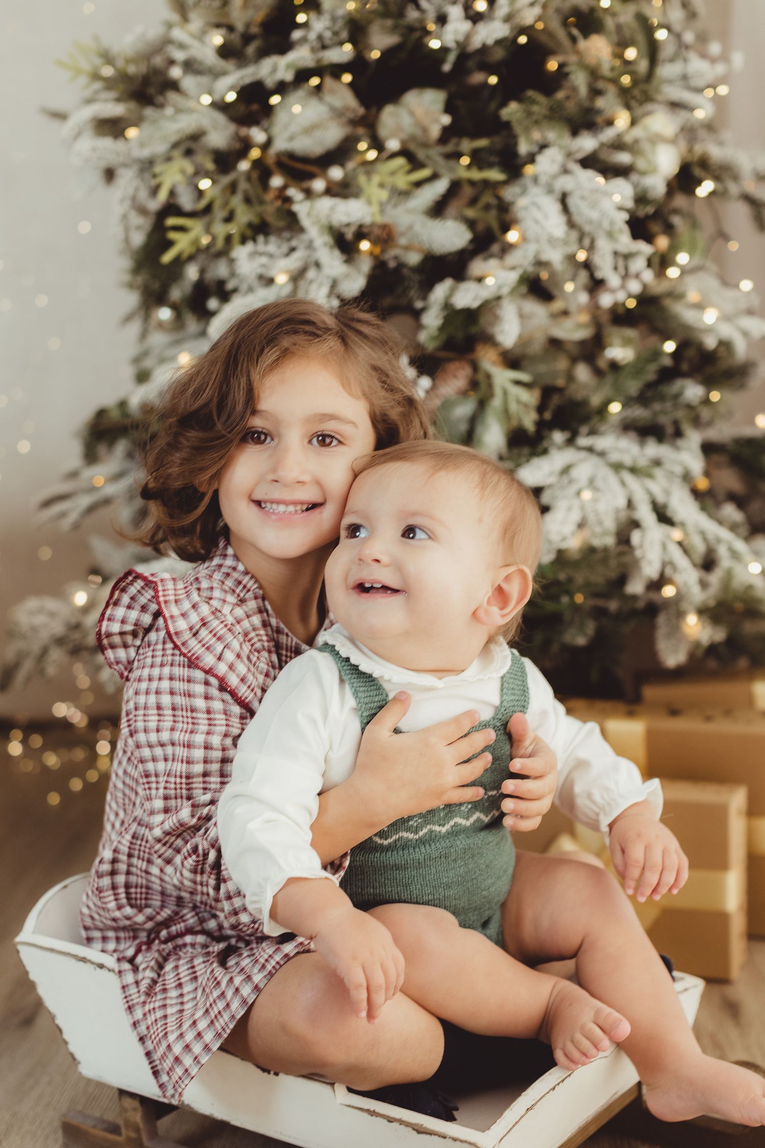 Two young children smiling in front of a decorated Christmas tree with warm glowing lights.