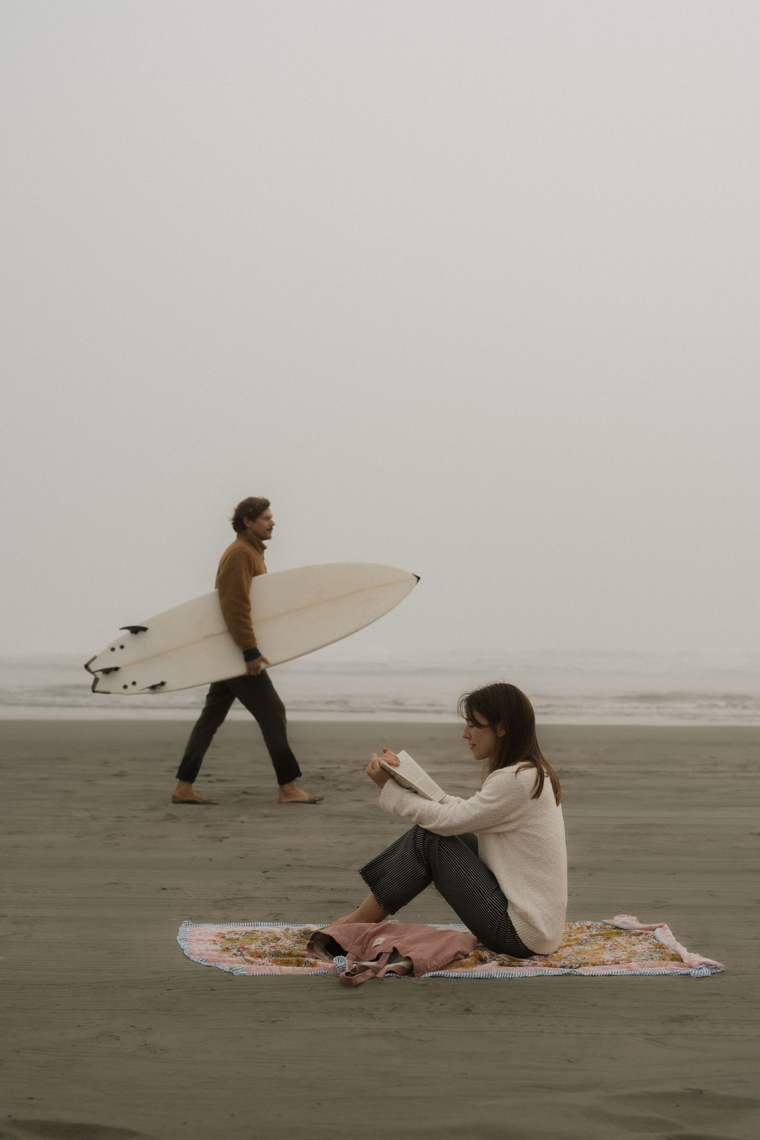 Couple on a foggy beach, woman sitting on blanket while man walks with white surfboard near ocean.