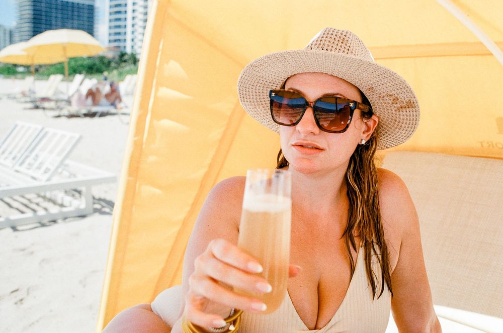 Woman in straw hat and sunglasses sipping a drink on a sunny beach.