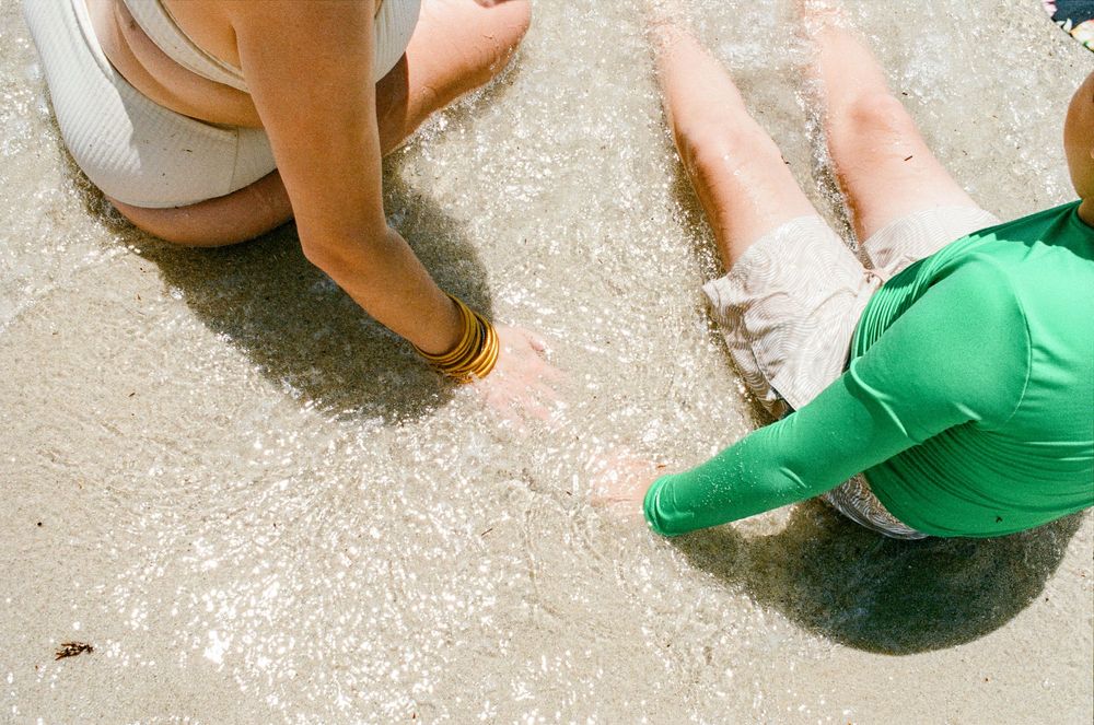 Two children playing in shallow beach waves, one wearing green top, with an adult nearby on sandy shore.