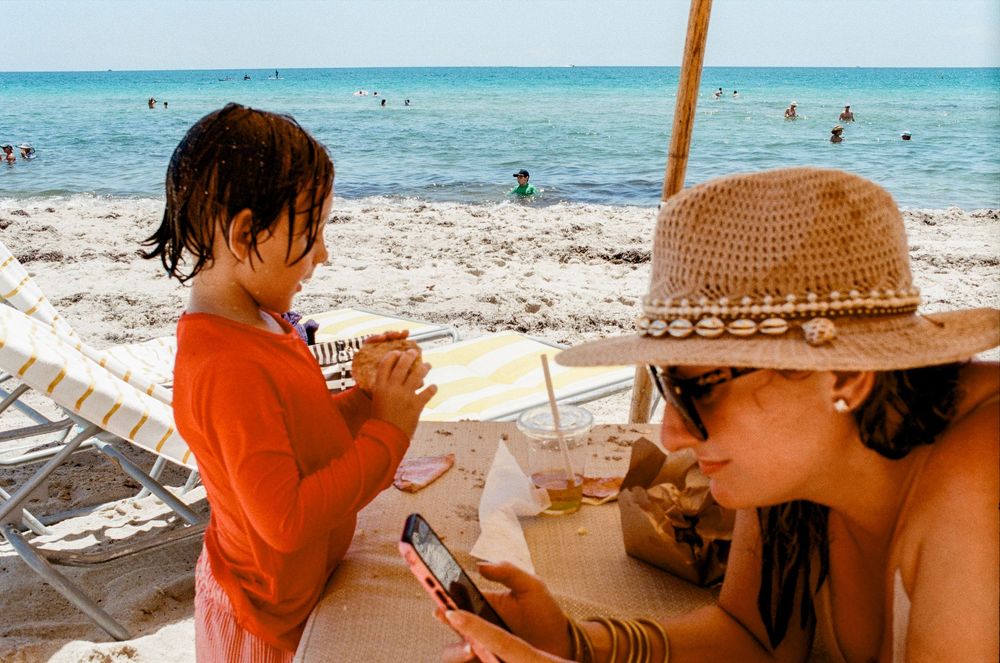 Child in orange shirt eating snack beside woman in straw hat using smartphone at a sunny beach.