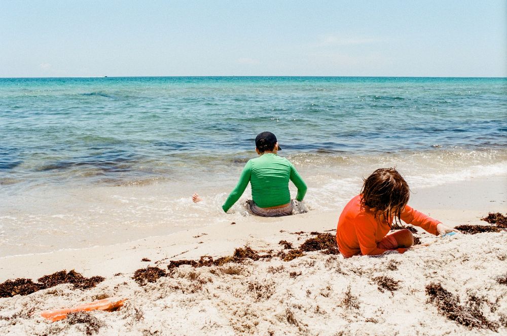 Two children sitting on a sandy beach shore, wearing colorful rash guards, gazing at calm turquoise ocean waters.