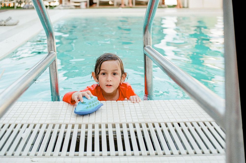 Young child in orange swimsuit clinging to pool ladder edge holding a blue kickboard during swim lessons.
