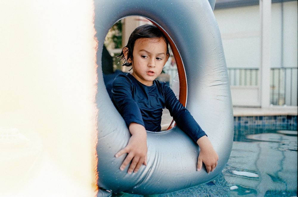 Young child sitting inside a large inflatable pool float ring near a swimming pool on a sunny day.