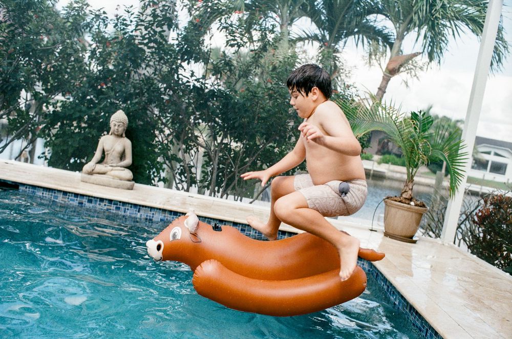 Boy jumping onto inflatable animal float in a backyard swimming pool with a Buddha statue and tropical plants.