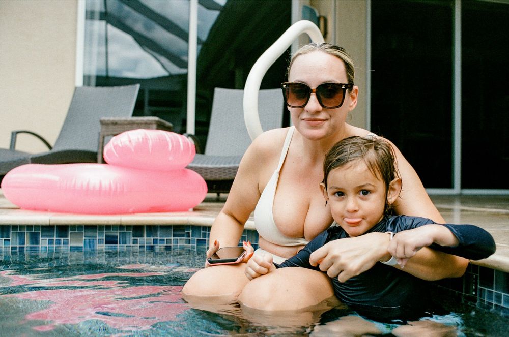 Woman and young child sitting at pool edge with pink inflatable ring float in background.