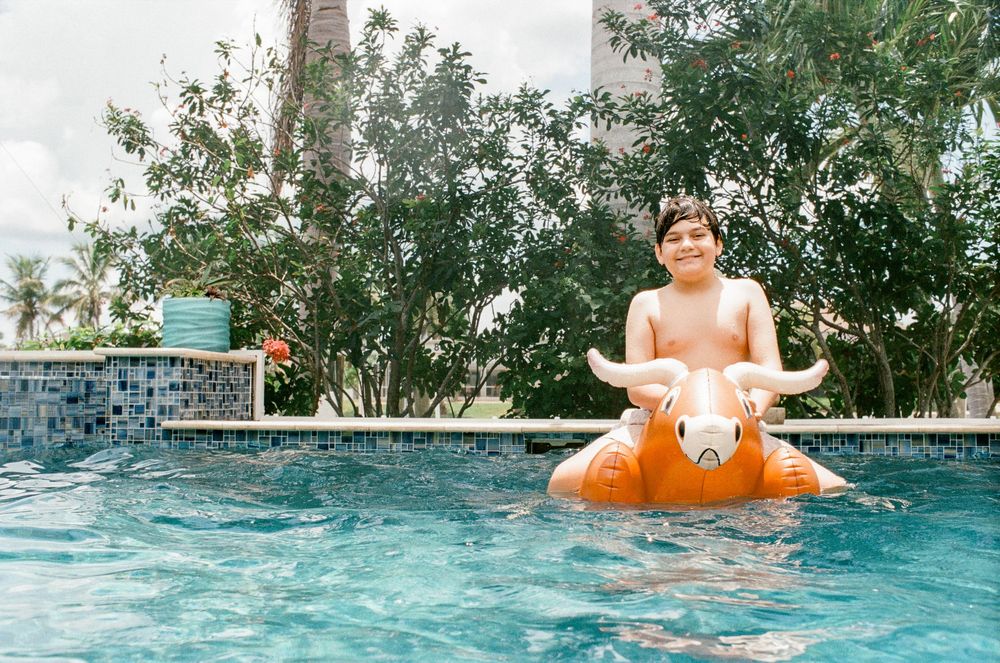Boy riding inflatable bull float in outdoor swimming pool surrounded by tropical trees.