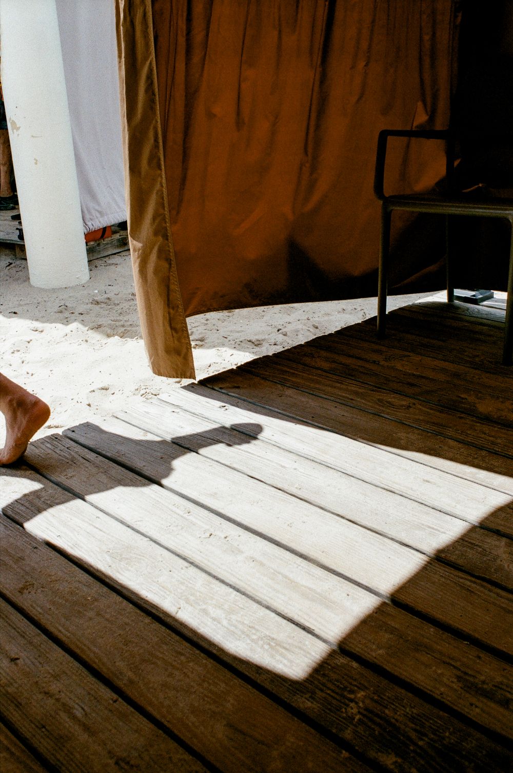 Shadow of a person cast on rustic wooden deck boards beside an orange curtain and chair in bright sunlight.