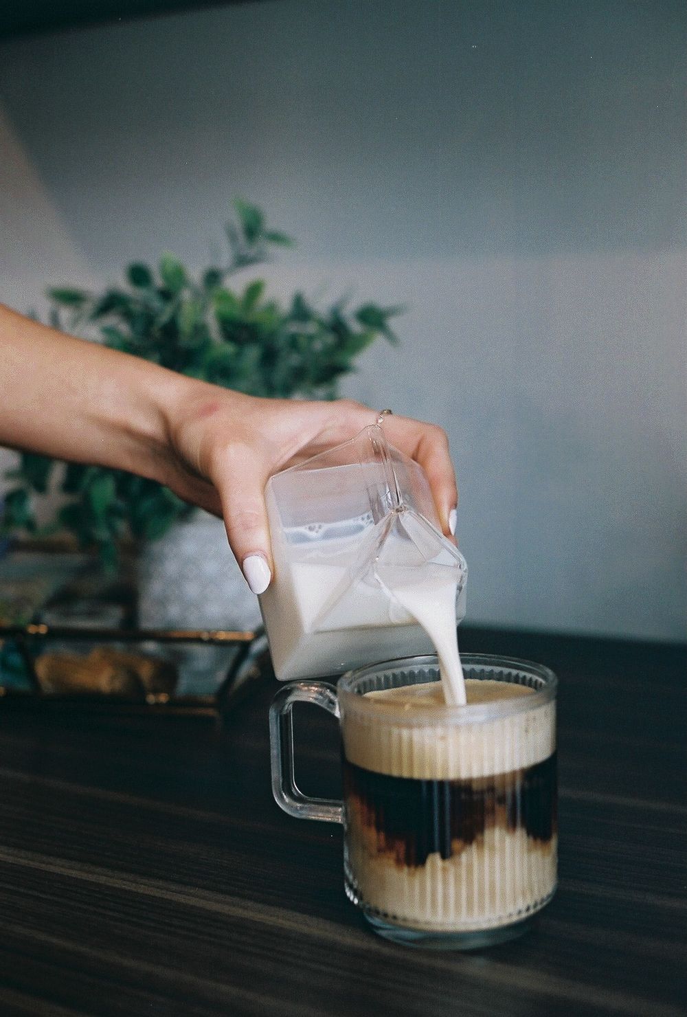 Hand pouring fresh milk into a glass mug of iced coffee on a wooden table with a plant in the background.