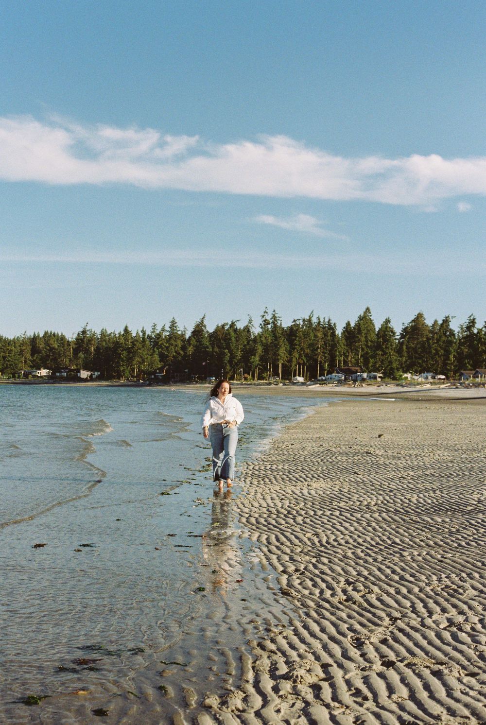 Woman in white shirt and jeans walking barefoot on a rippled sandy beach with trees in background.