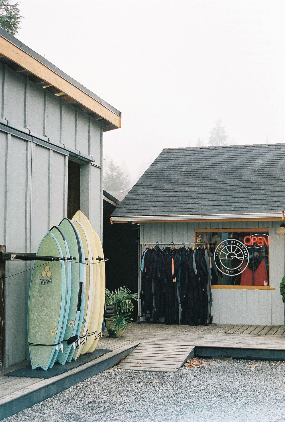Surf shop exterior with colorful surfboards leaning against wall and wetsuits displayed near open sign on foggy day.