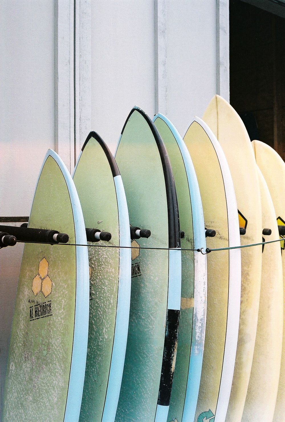 Multiple colorful surfboards stored upright in a rack against a white wall, displaying shades of blue, green, and yellow.
