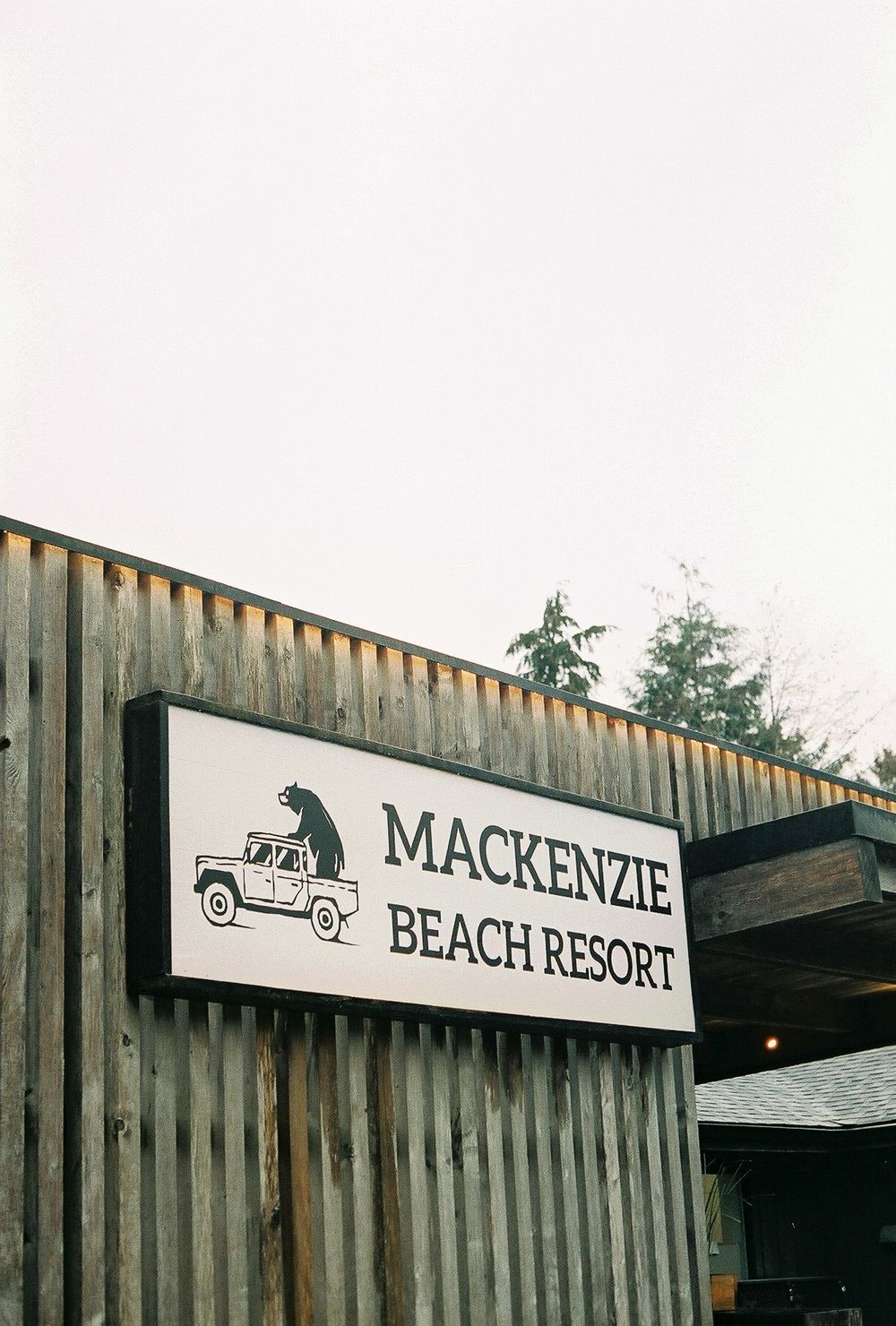 Sign for Mackenzie Beach Resort on a rustic wooden building with a bear on a truck logo, surrounded by trees.