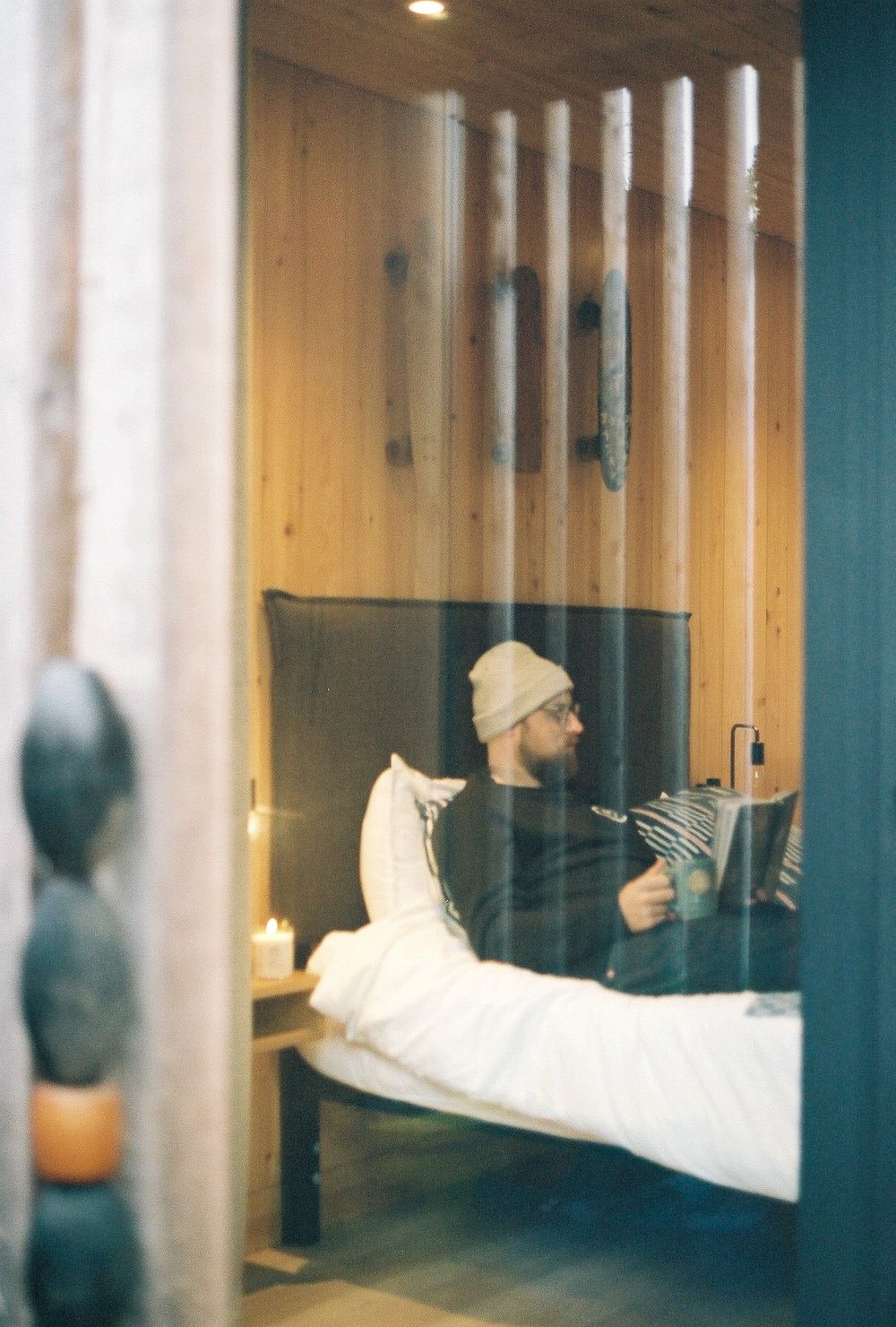 Man in beanie hat reading book on bed with candle, viewed through glass window in cozy wood-paneled room.