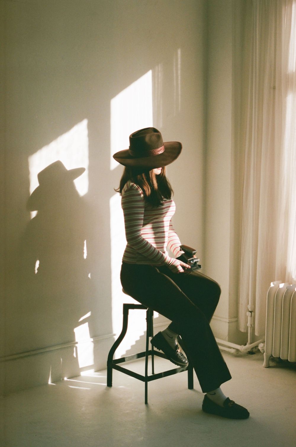 Woman in wide-brim hat and striped top sits on stool in sunlit room with dramatic shadows on white wall.