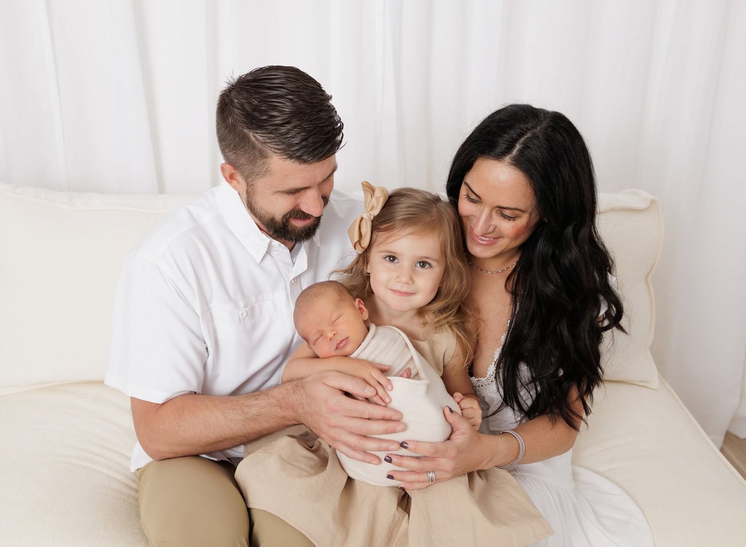 Happy family of four with newborn baby, toddler girl, mom and dad sitting together on a cream couch.