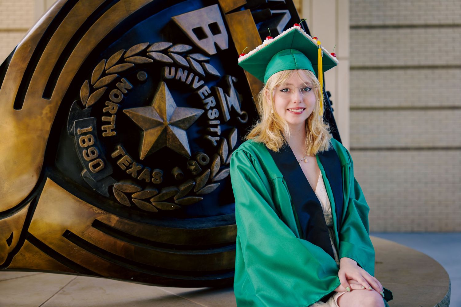 Female graduate in green cap and gown posing beside University of North Texas seal.