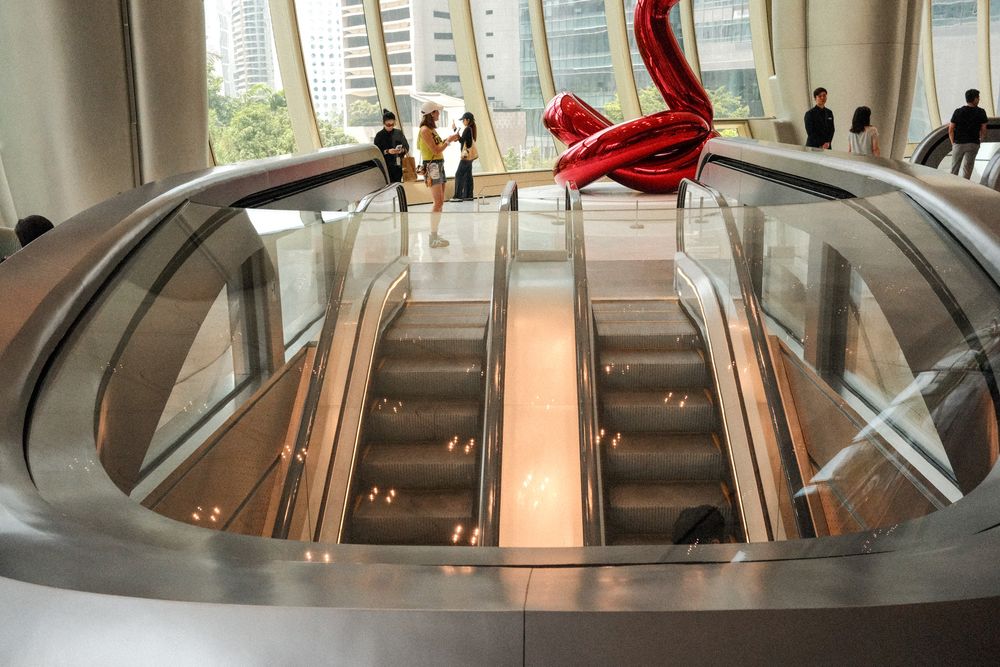Circular escalator descending into a modern atrium with a red sculpture and city views.