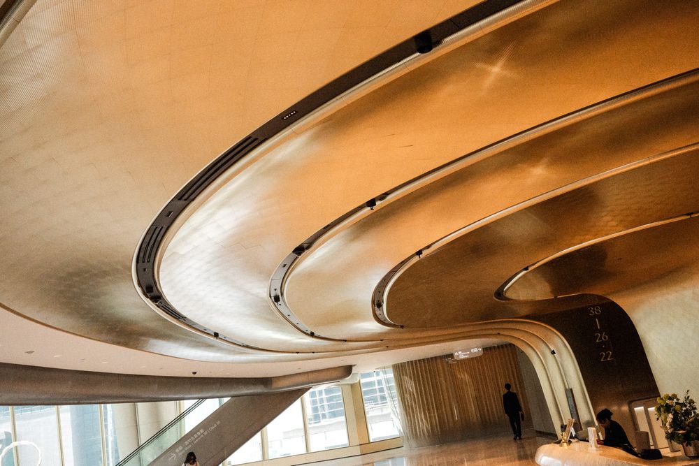 Modern building interior featuring sweeping curved golden ceiling layers with dramatic architectural design and a silhouetted figure below.