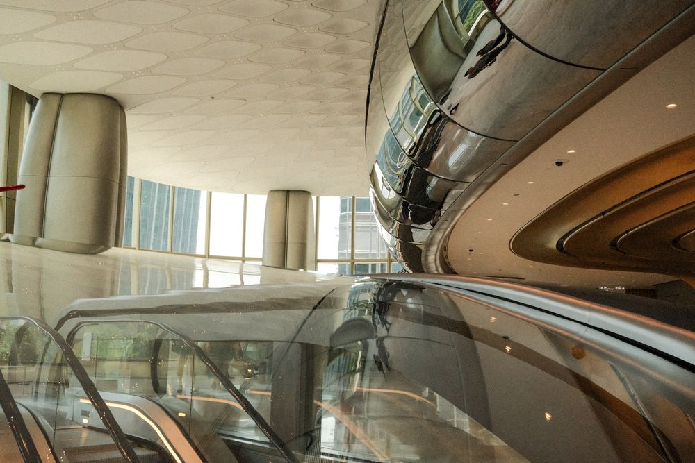 Modern mall interior with escalators, reflective curved walls, tall columns, and large windows in a luxury shopping center.