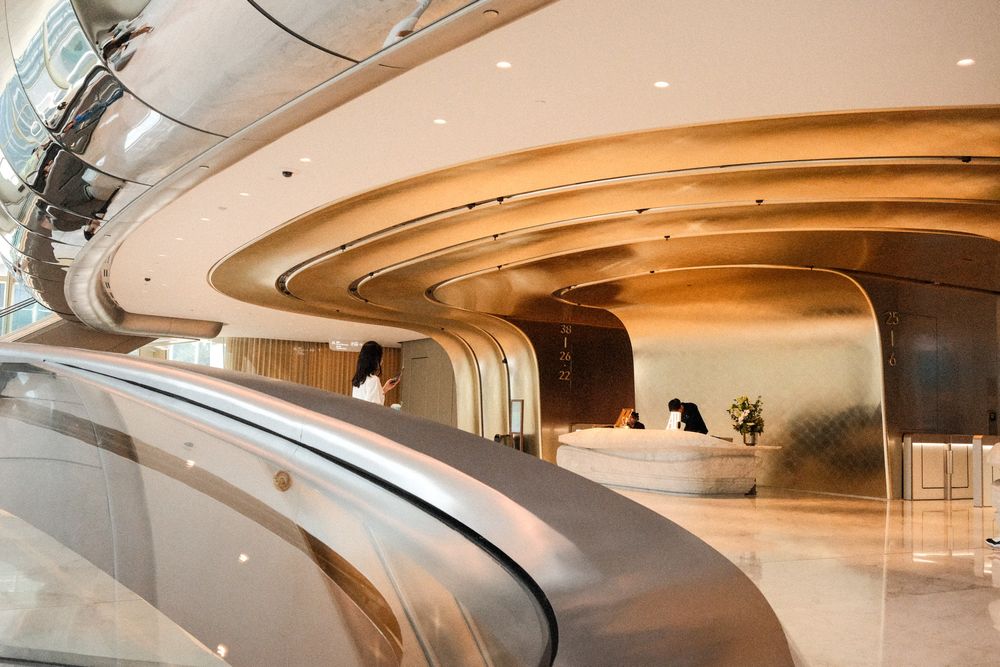 Futuristic luxury hotel lobby with sweeping curved gold metallic ceilings, escalators, and reception desk in modern architecture.