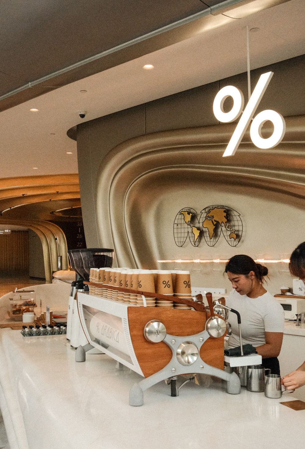 Barista working at % Arabica coffee shop with espresso machine, world map decor, and glowing % sign.