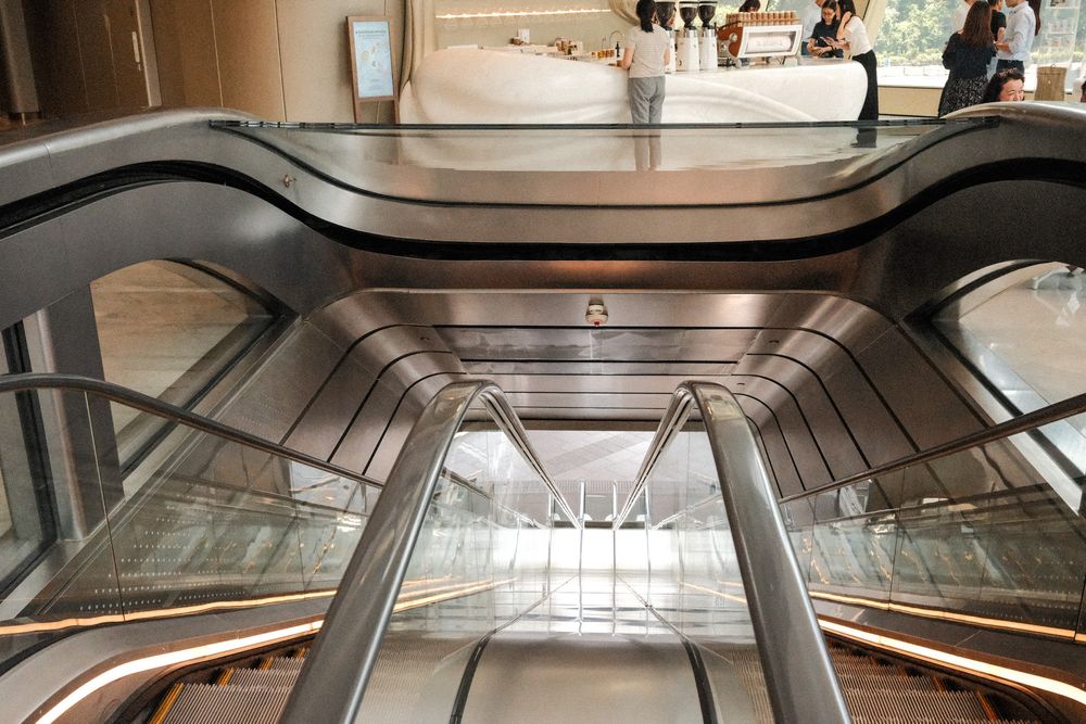 Modern escalators in a shopping mall with sleek silver railings and glass panels viewed from above.