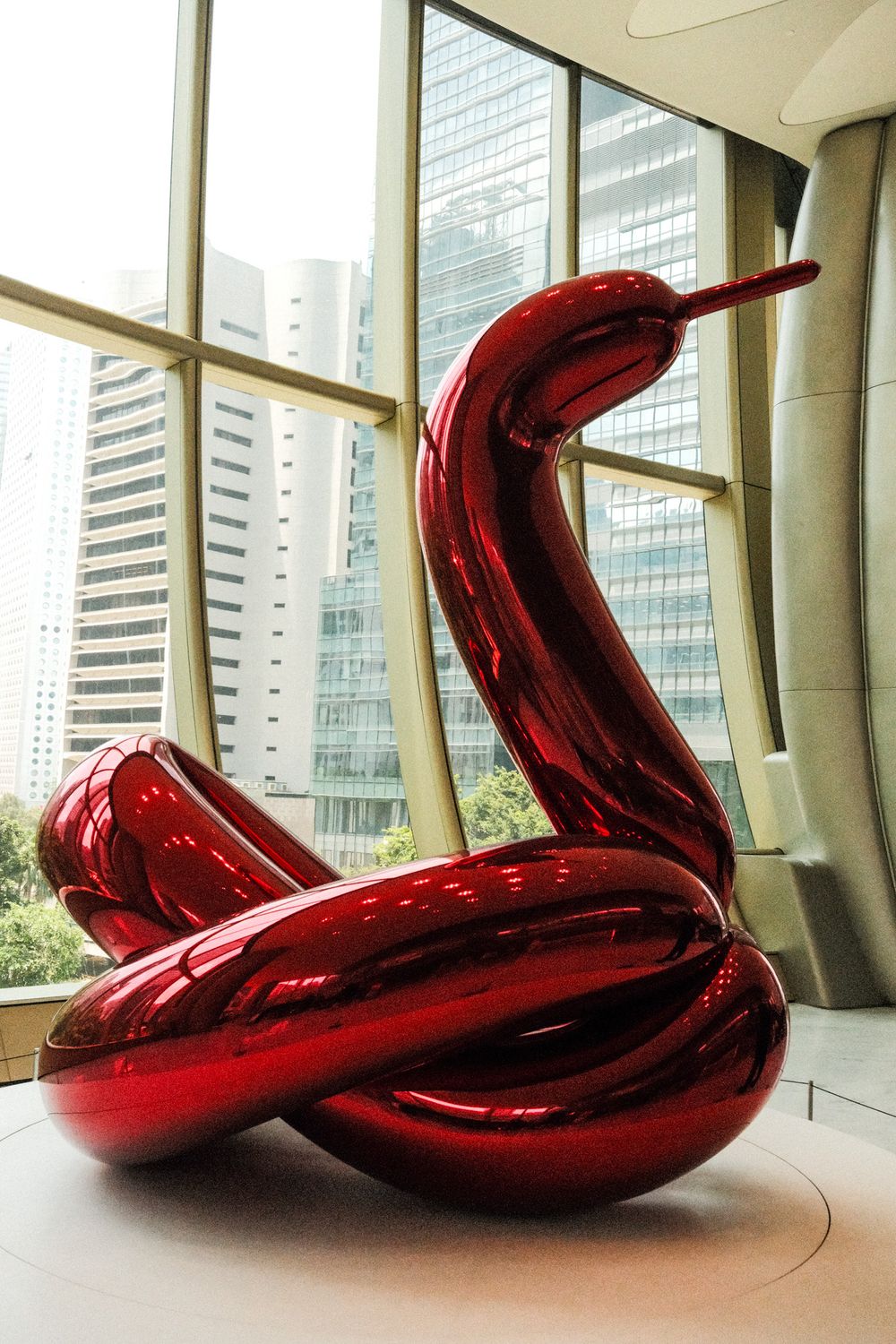 Red metallic balloon swan sculpture displayed indoors with city skyscrapers visible through large windows.