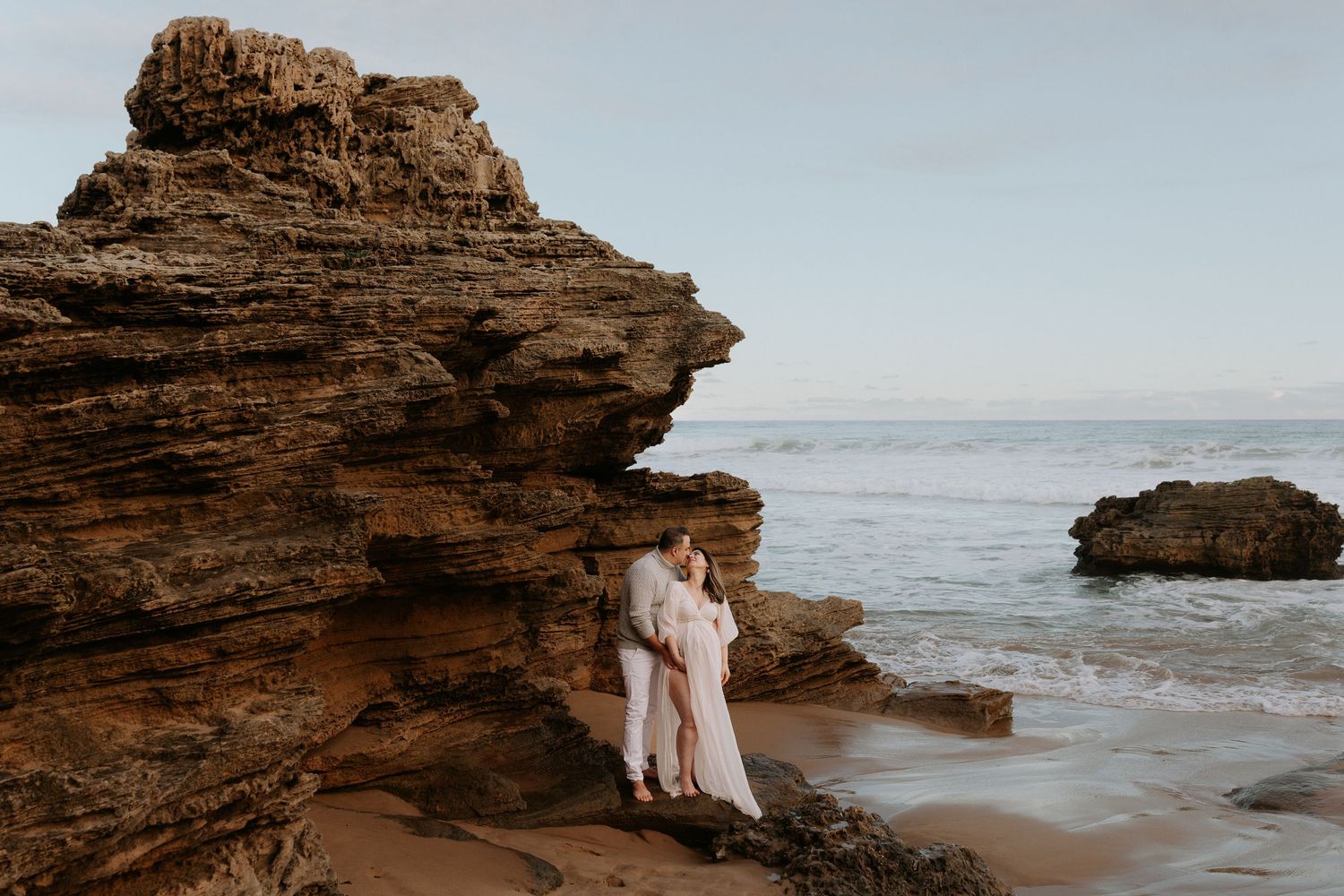 Couple embracing on rocky beach shore with ocean waves and dramatic sandstone cliffs in background.