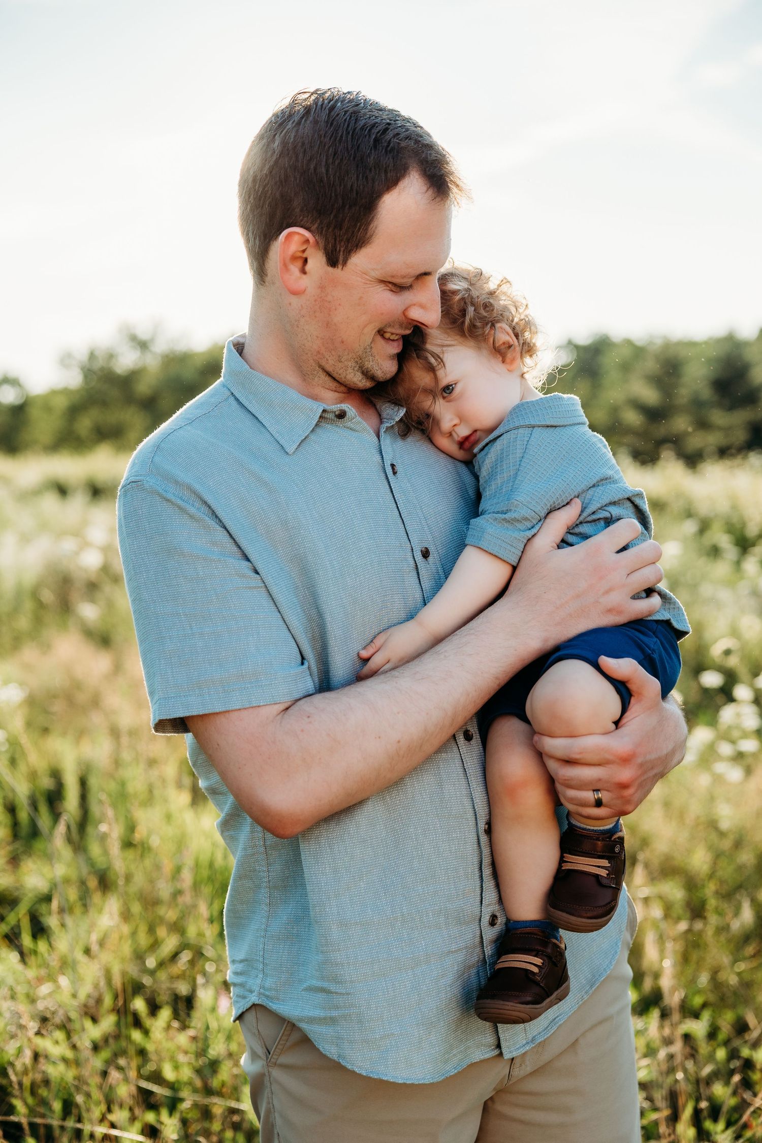 A father tenderly holds his young toddler in a sunny wildflower field, both wearing blue shirts.