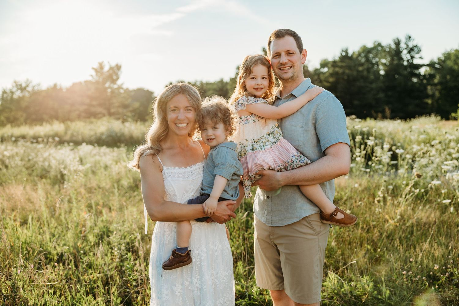 Family of four smiling in a sunny wildflower meadow, parents holding young children during a golden hour outdoor portrait.