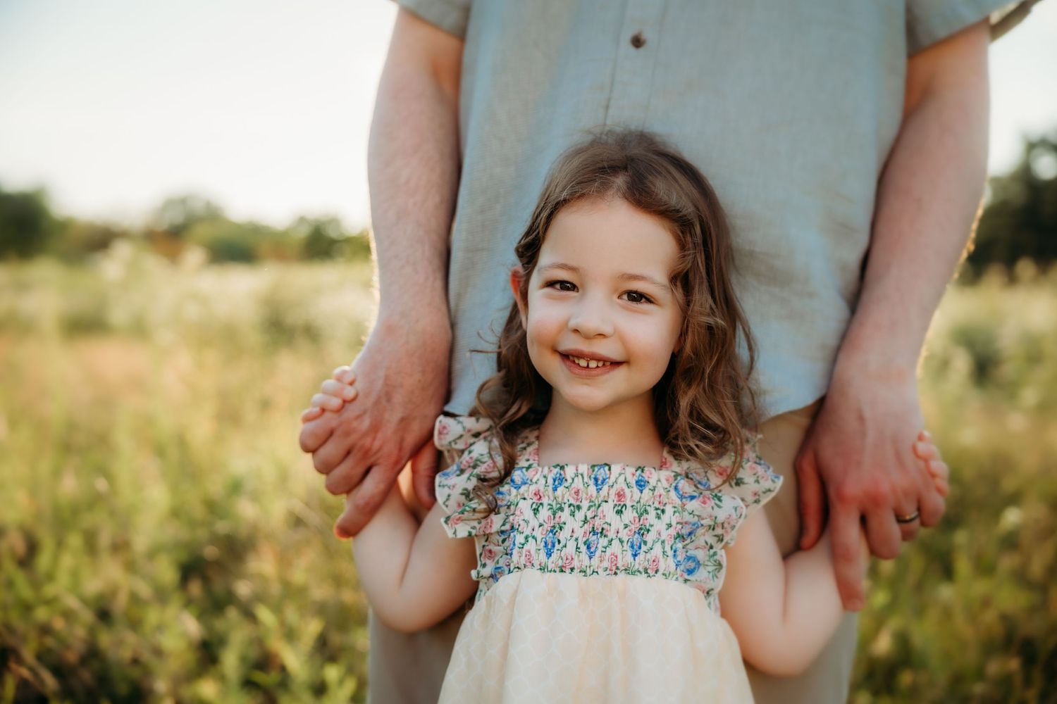 A smiling little girl in a floral dress holds an adult's hands in a sunny green field.