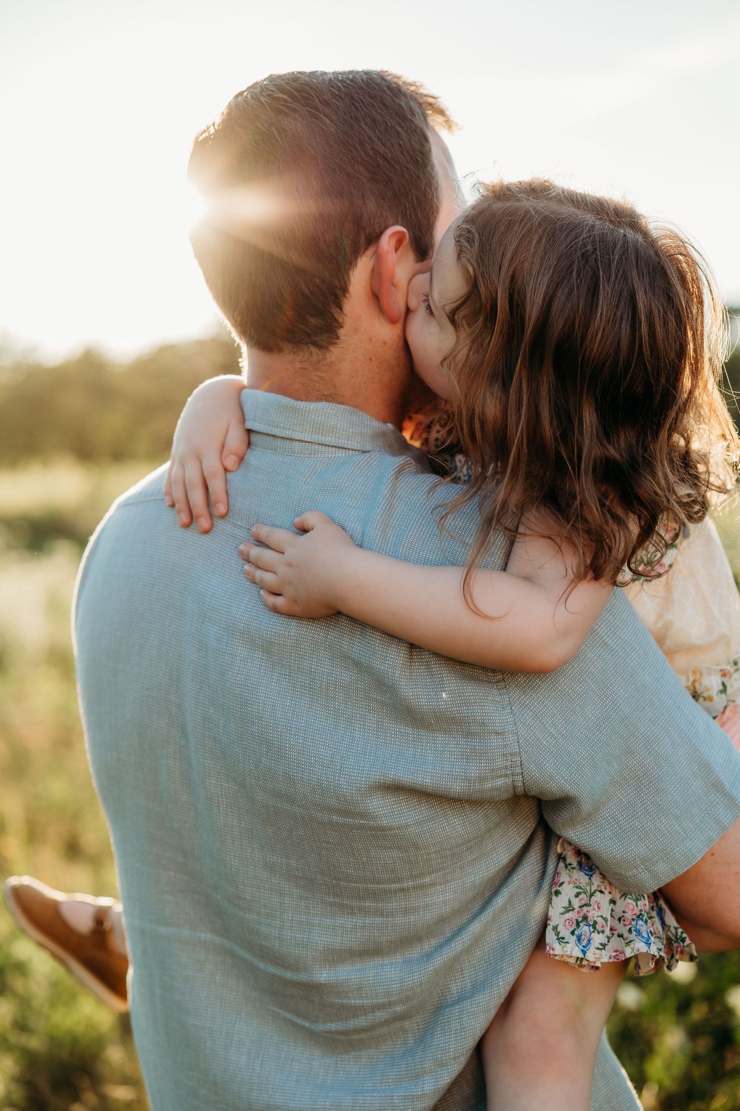 A father holds his young daughter in a warm sunset embrace as she kisses his cheek in an outdoor field.