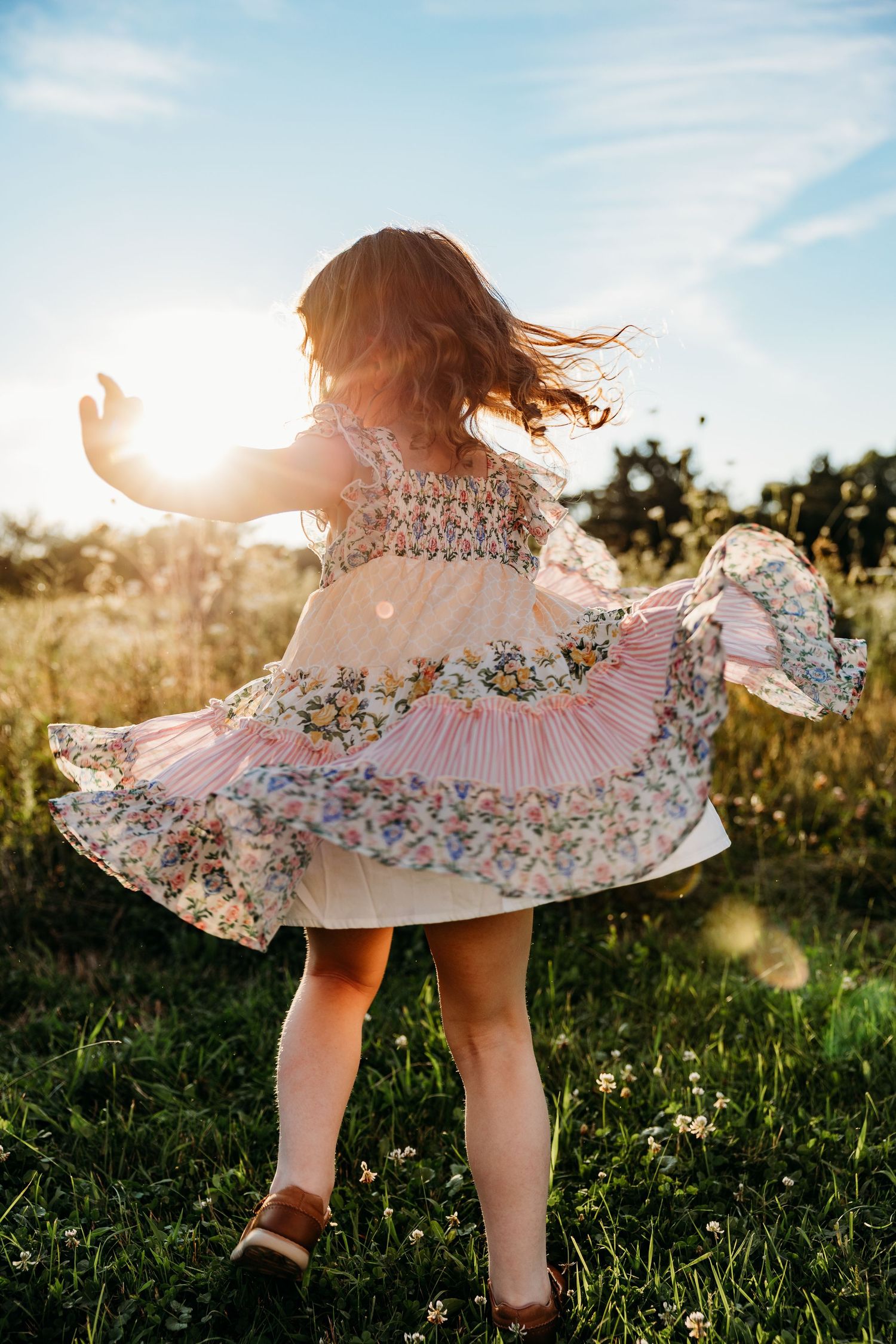 Young girl twirling in a floral dress in a sunny meadow, hair flowing, backlit by golden sunlight.