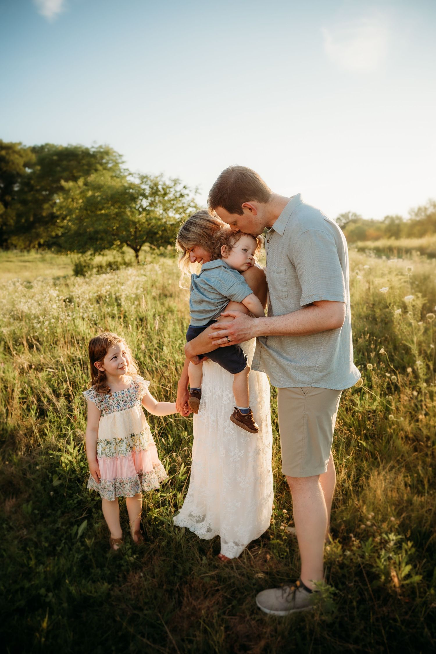 Family of four enjoying a sunny outdoor photo session in a green meadow, father kissing baby while daughter smiles.