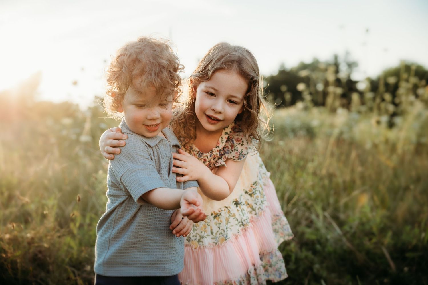 Two young children hugging outdoors in a sunny field, a curly-haired boy and a smiling girl in a pink dress.
