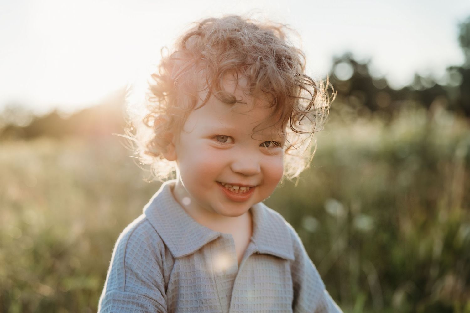 Smiling toddler with curly hair outdoors in golden sunlight, wearing a light blue shirt in a sunny meadow.