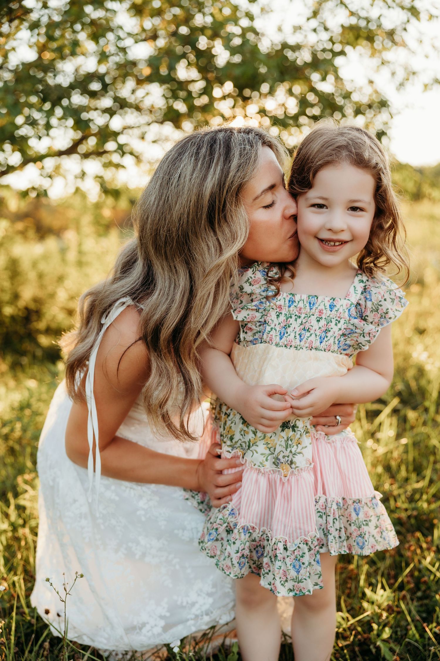 A woman in a white dress kisses a smiling little girl in a floral dress outdoors in a sunny green field.