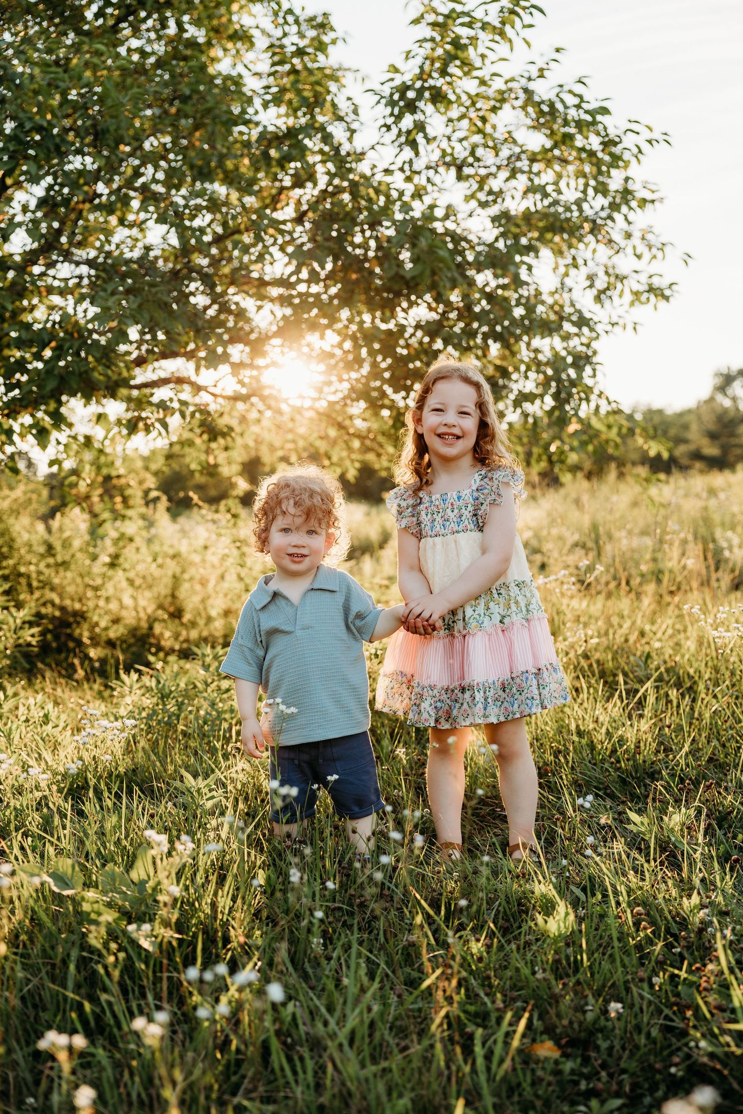 Two young children smiling in a sunny meadow, girl in floral dress and boy in blue shirt, golden hour light.