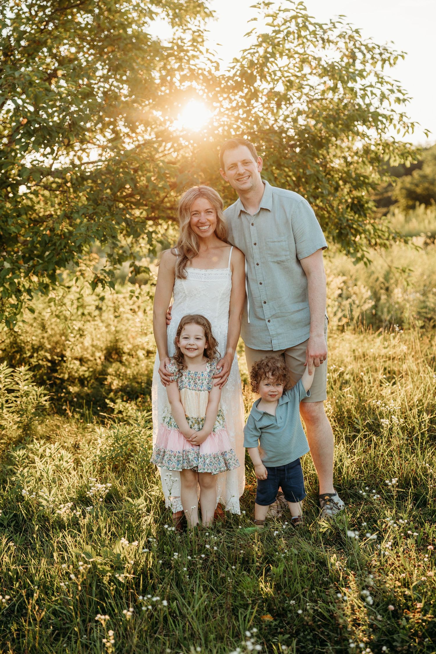 Family of four posing outdoors at golden hour, parents with two young children in a sunlit meadow.