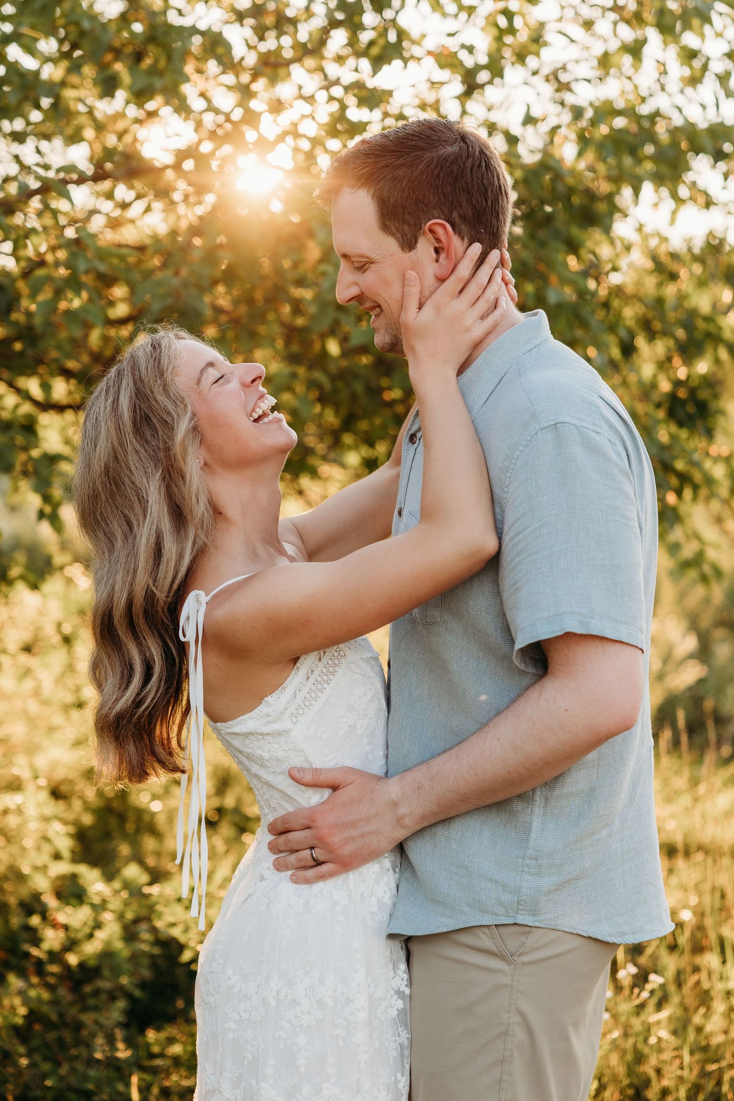 A laughing couple embraces outdoors at golden hour, woman in white lace dress, man in blue shirt, with sunlit trees behind them.
