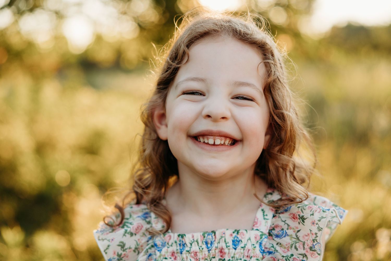 A joyful young girl with curly hair smiles brightly outdoors in a floral dress, bathed in warm golden sunlight.