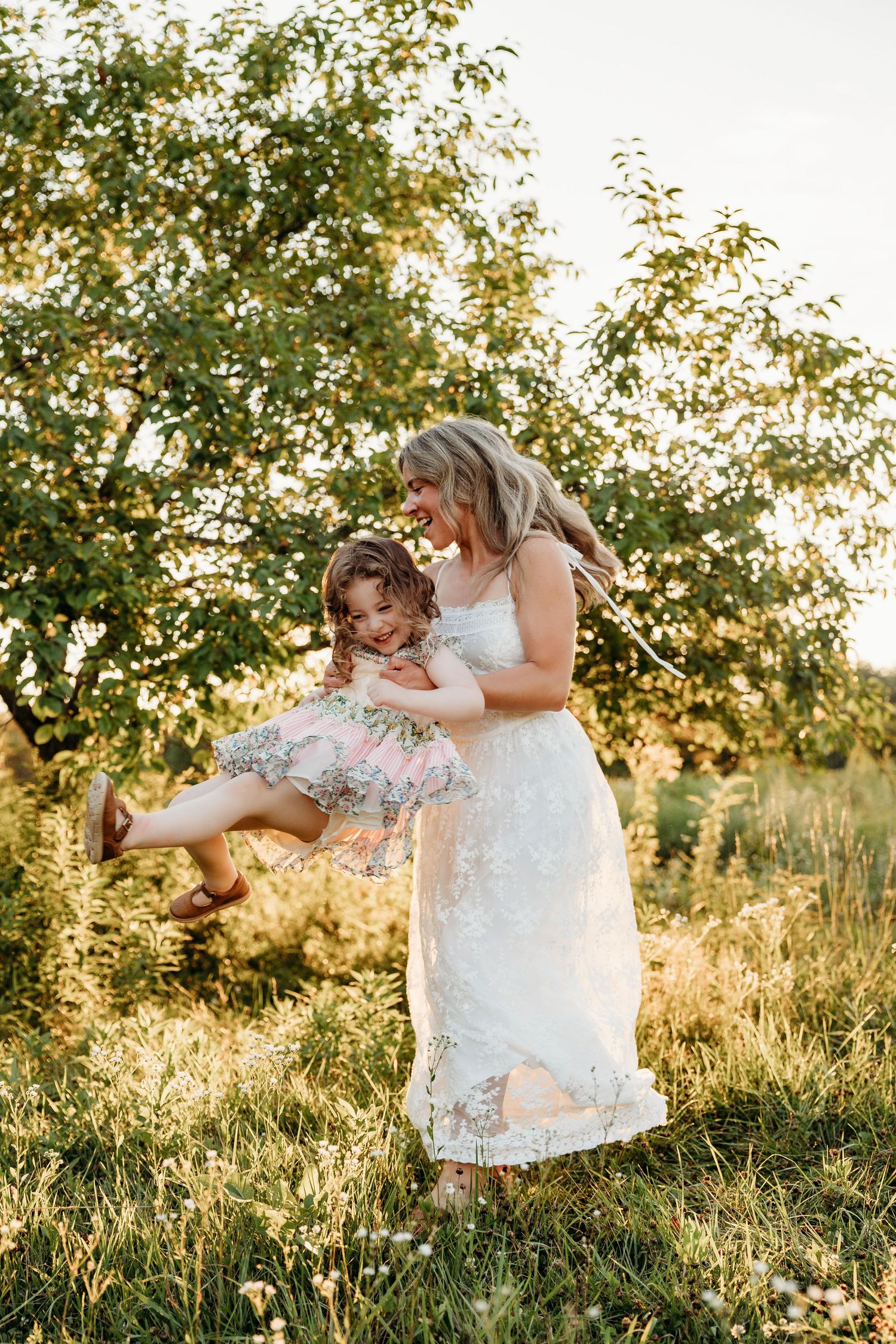 A smiling mother in a white lace dress swings her laughing daughter outdoors in a sunlit green garden.