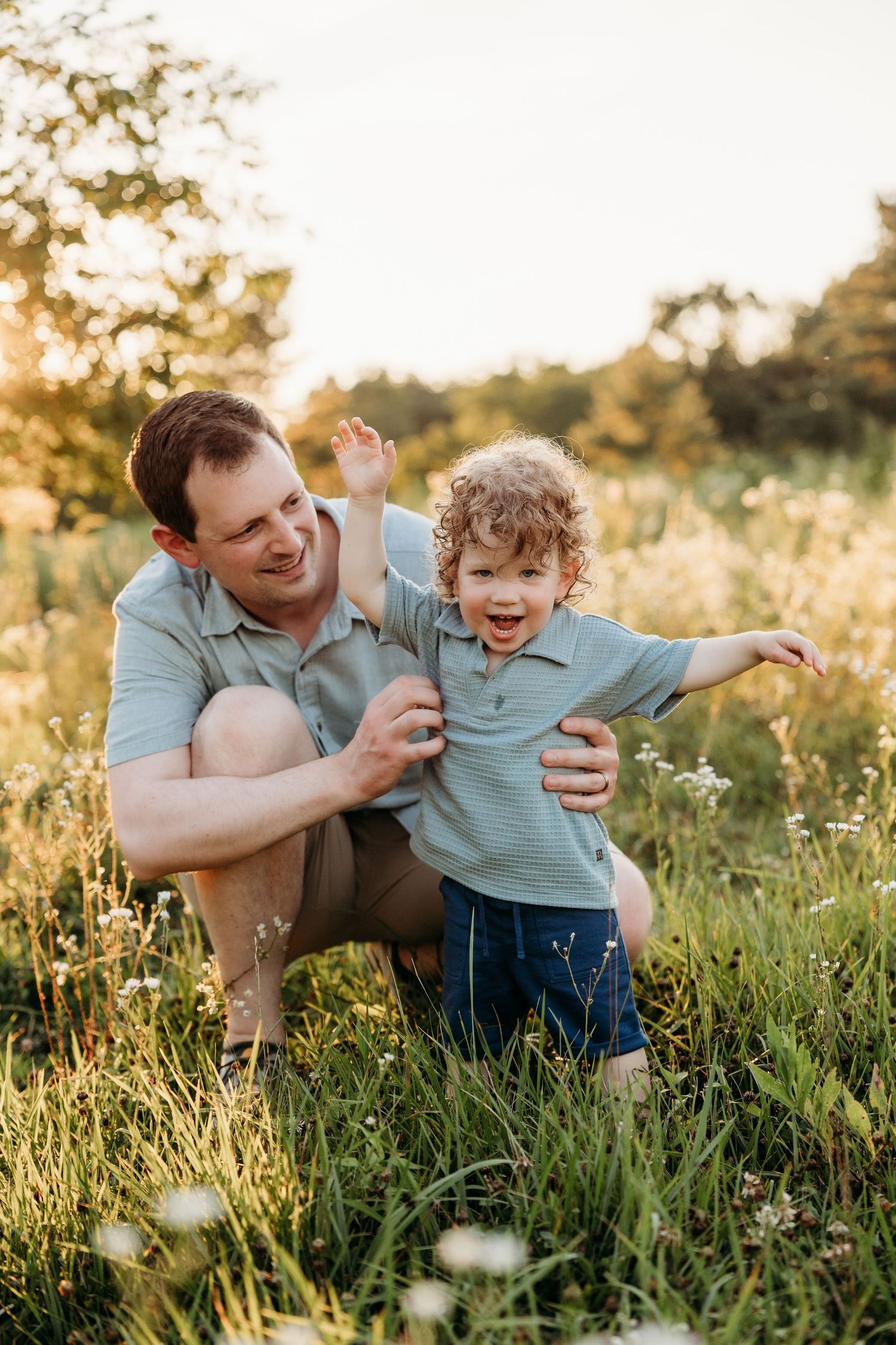 A father crouches in a sunny meadow, joyfully holding his curly-haired toddler son with arms outstretched.