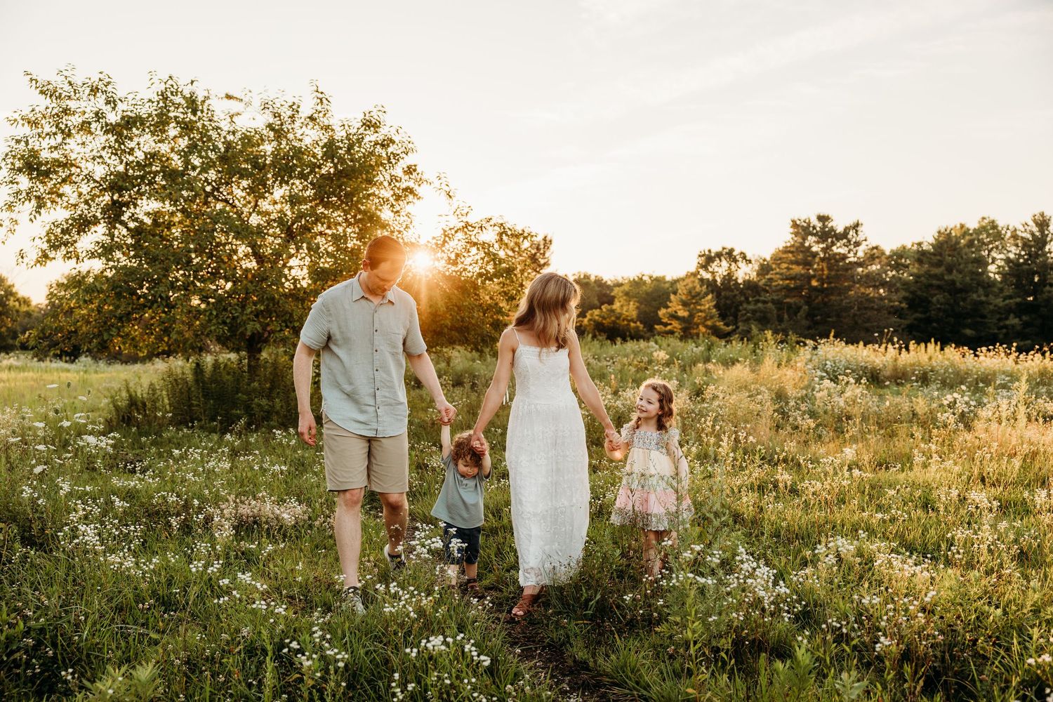 Family walking through a sunny wildflower meadow at golden hour, parents holding hands with two young children.